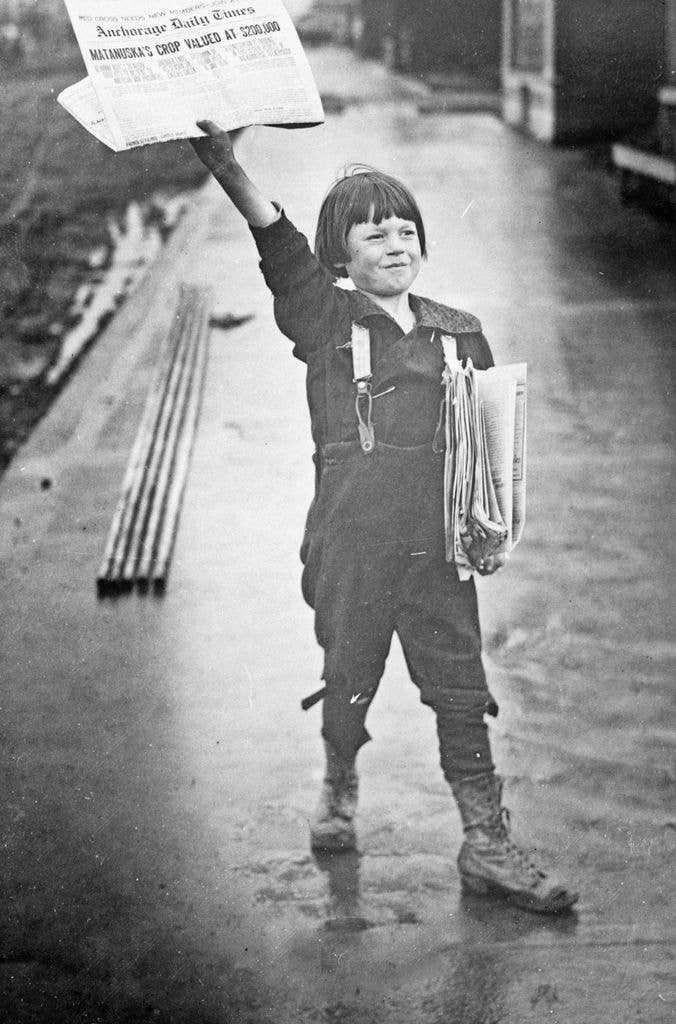 A small boy holding up a newspaper with a pile under his arm.