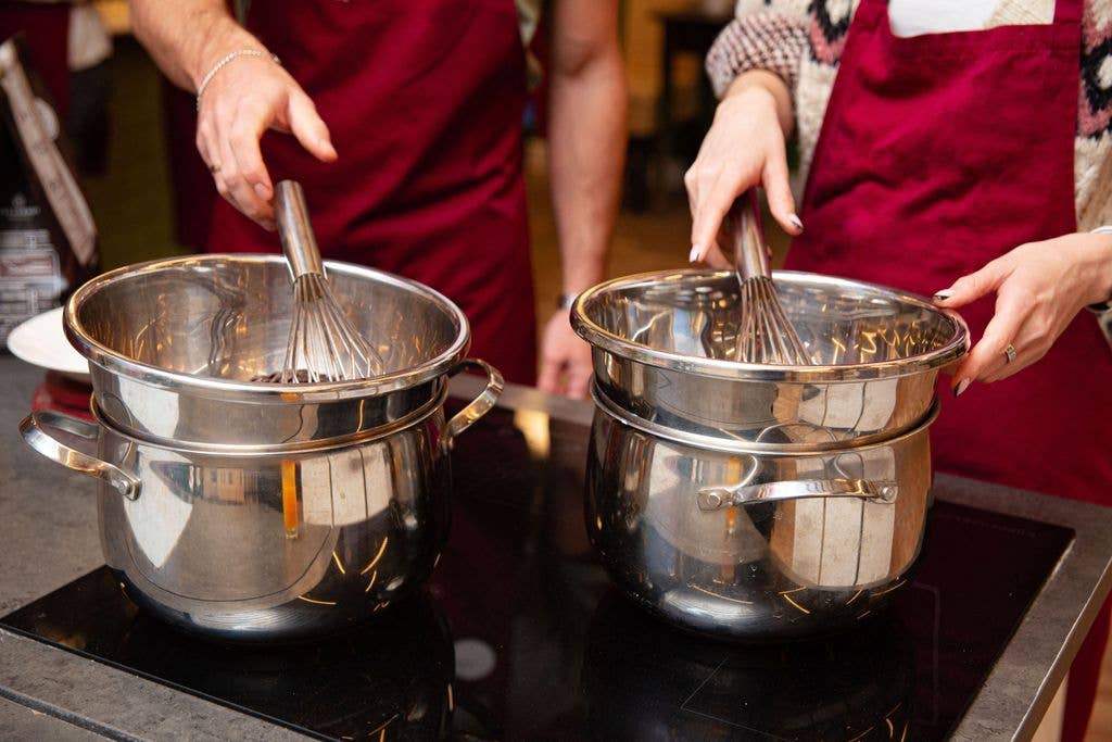 Two people wearing burgundy aprons each stir a stainless steel double-boiler pot containing melting chocolate, using whisks, at a cooking class or workshop.