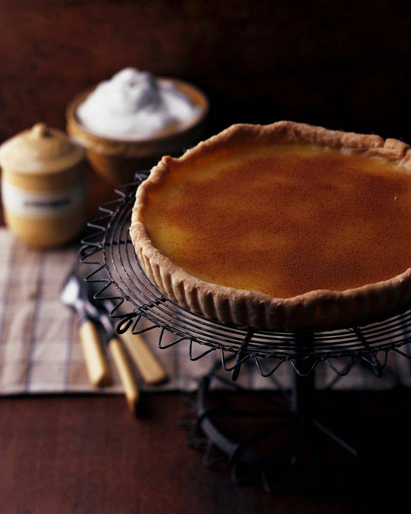 A cinnamon-dusted vinegar pie in a fluted pastry crust sits on a wire cooling rack, with a bowl of whipped cream and a jar in the background.
