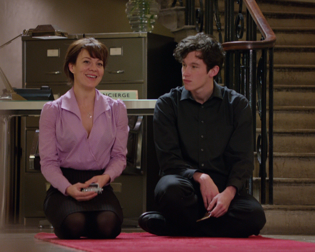 A woman in a pink blouse and a man in a black shirt sit on a red rug, smiling.