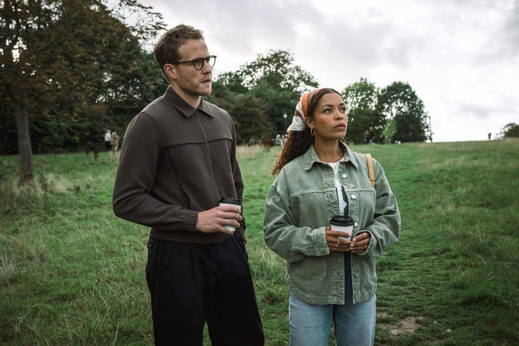 Man and woman standing in a grassy field, both holding coffee cups.