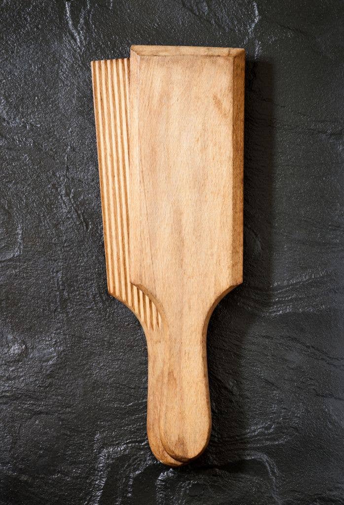 A pale wooden kitchen paddle with a grooved, ridged texture and rounded handle, photographed against a dark grey slate surface.