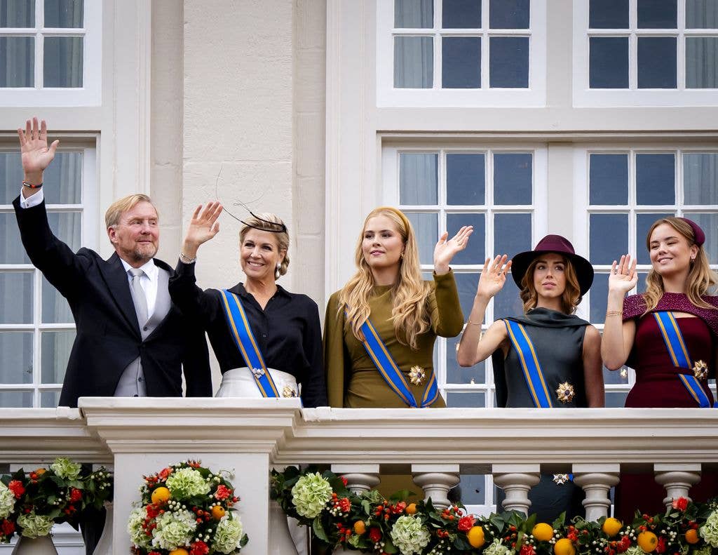Queen aáxima, Willem-Alexander, and their daughters wave from a balcony adorned with flowers.