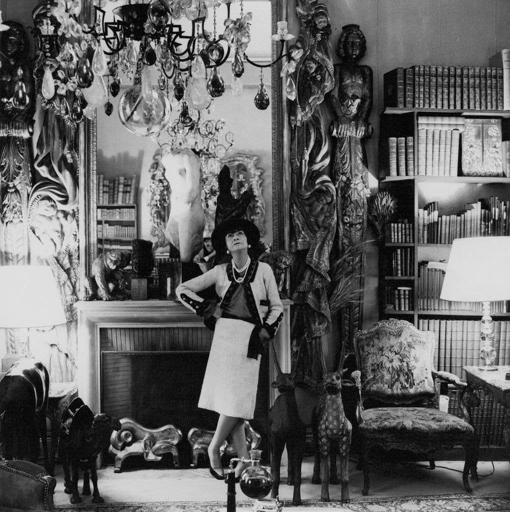 French fashion designer, Coco Chanel, standing, in her Paris apartment, with her back to a mantle fireplace, with a large mirror above, looking up at a chandelier.