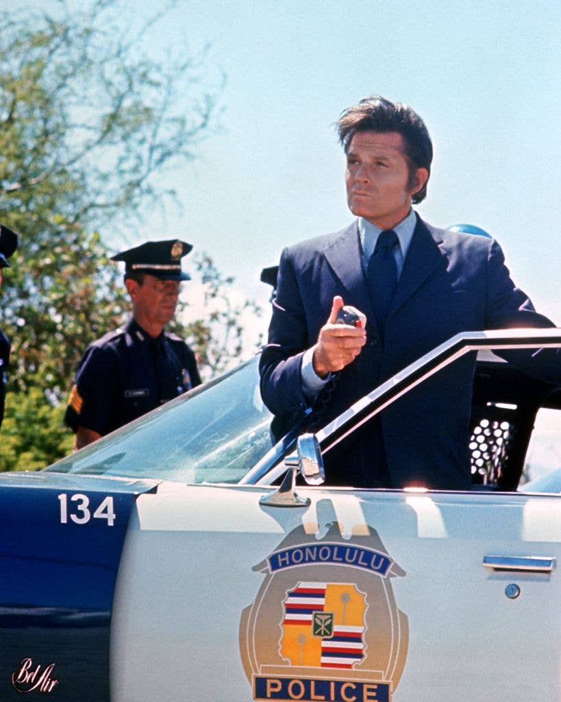A man in a dark suit holds a car radio microphone while standing at the open door of a Honolulu police car, with a uniformed officer looking on behind him.