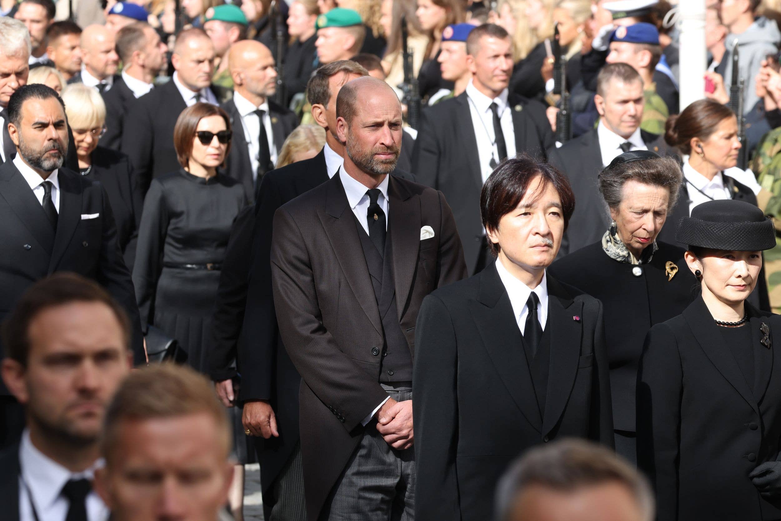 Prince William stands among a crowd in formal attire, looking solemn.