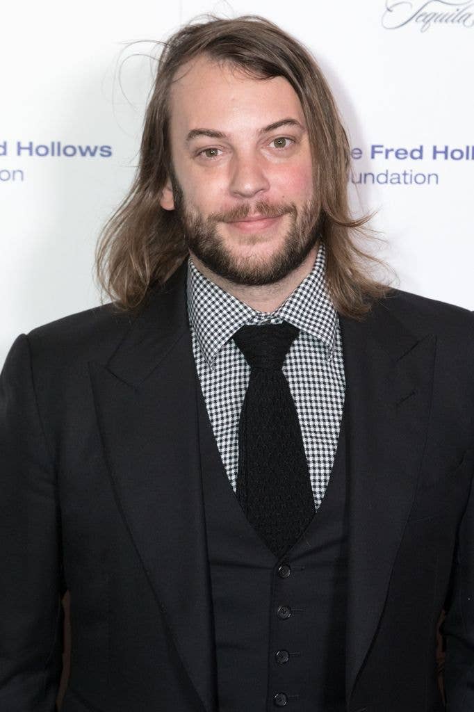 Angus Sutherland, with long hair and a beard, wears a black three-piece suit and checked shirt as he poses at a step-and-repeat for the Fred Hollows Foundation gala.
