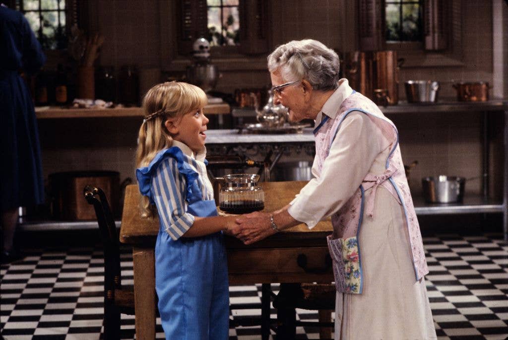 A young girl in a blue striped dress shakes hands with an older woman wearing an apron in a period kitchen with checkerboard floor.