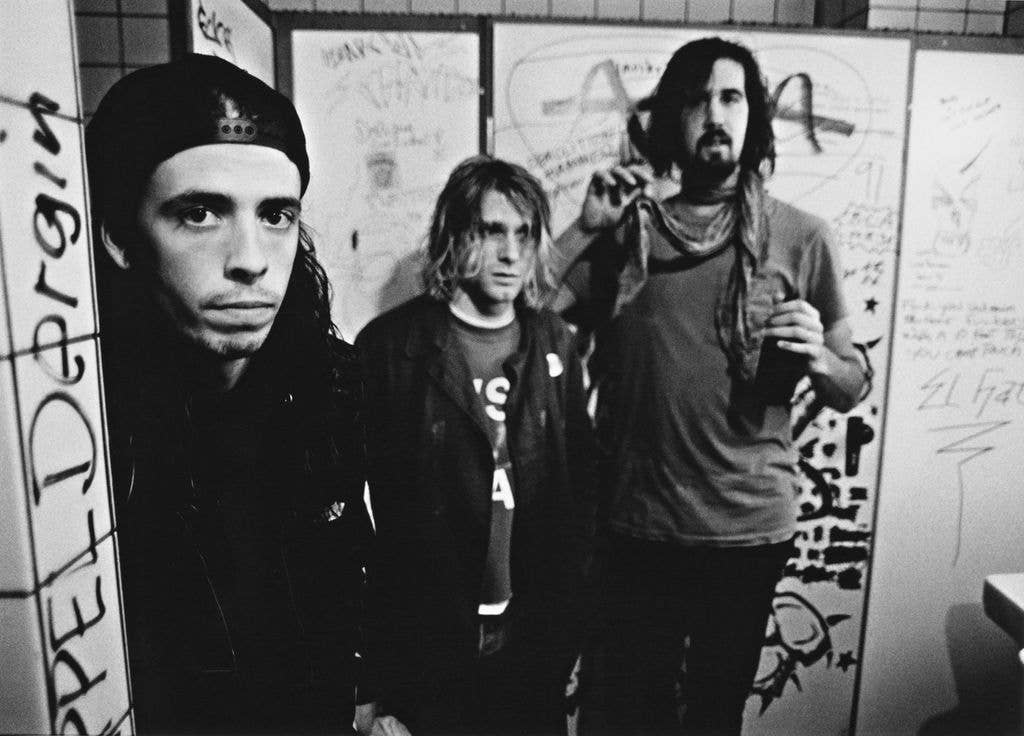 Black and white photo of Dave Grohl, Kurt Cobain and Krist Novoselic standing backstage against a graffiti-covered wall in Frankfurt in 1991.