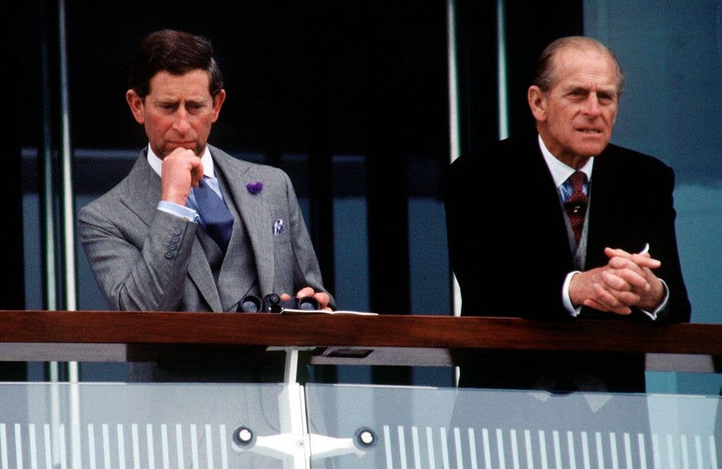 The Prince of Wales in a grey suit stands beside Prince Philip in a black suit, both on a balcony.