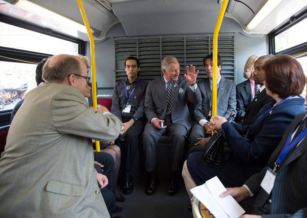 Charles, Prince of Wales, in a gray suit and striped tie, waves while seated on a bus with other formally dressed individuals.