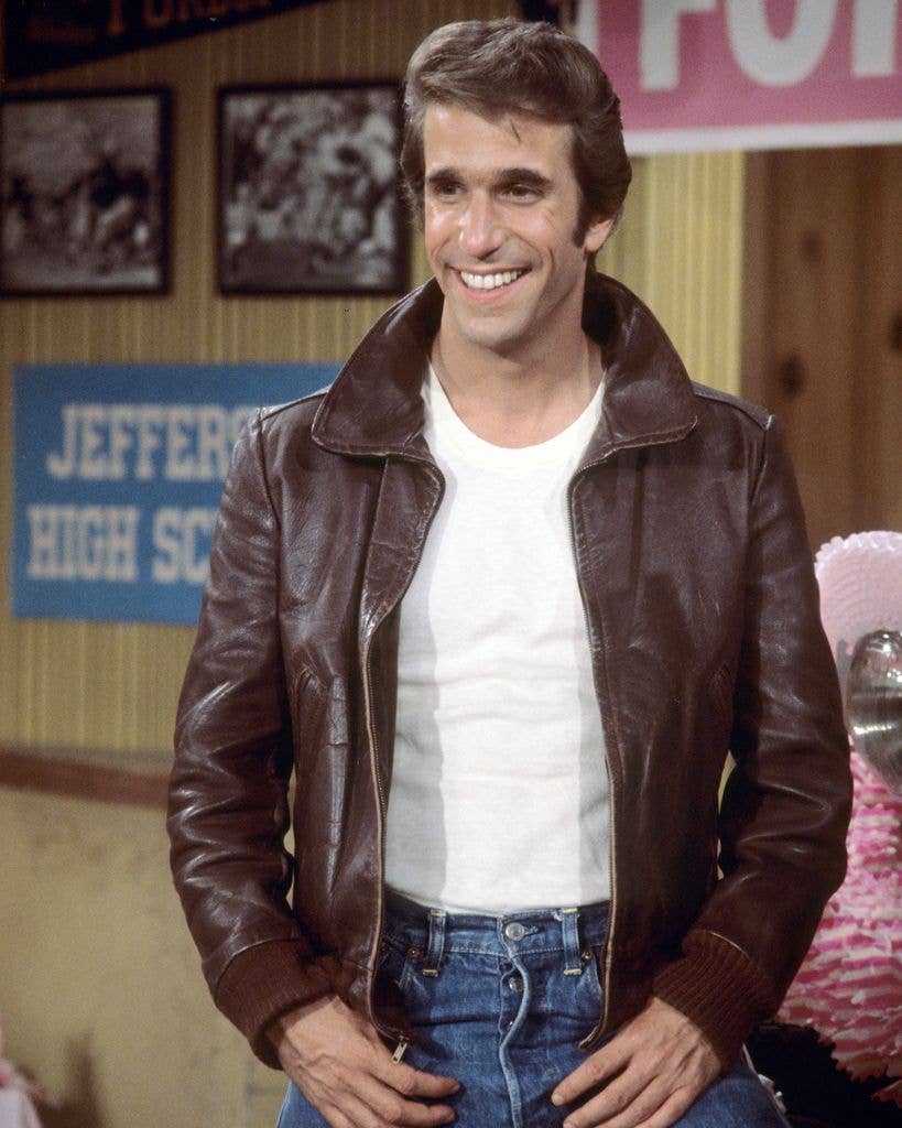 Henry Winkler, smiling broadly, wears a brown leather jacket over a white t-shirt in a 1970s American high school set.