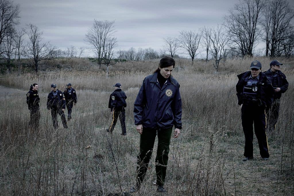 Michelle Monaghan, in a blue jacket, stands among law enforcement officers in a field.