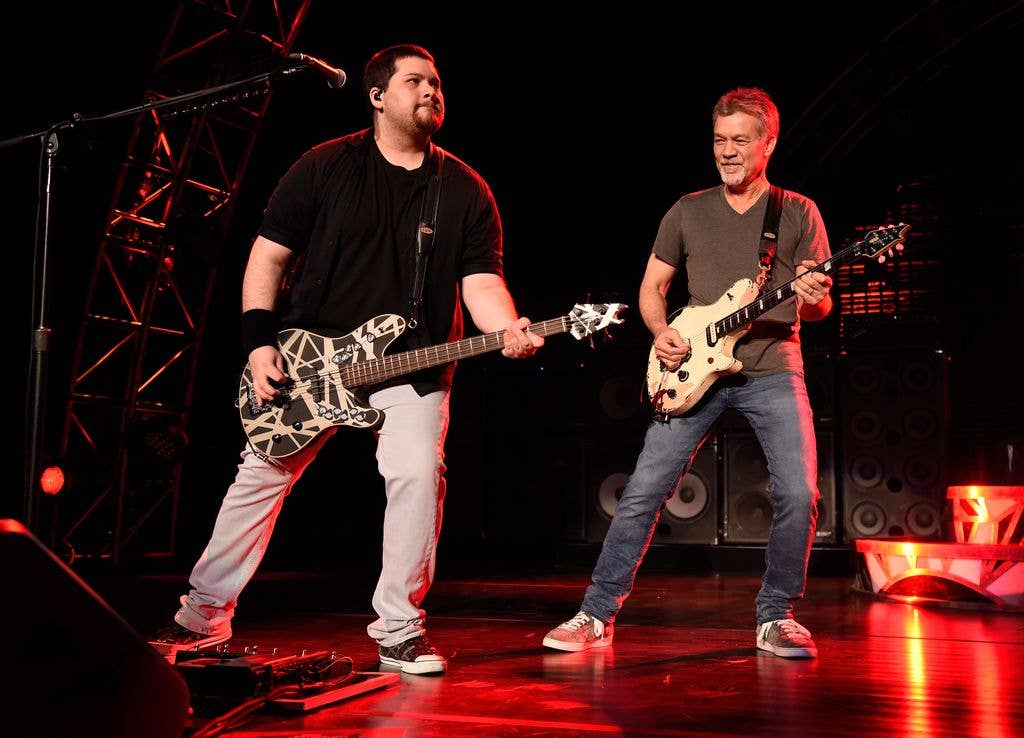 Two guitarists perform on a stage lit with red lighting, one on bass and one on lead guitar, both wearing striped instrument straps.