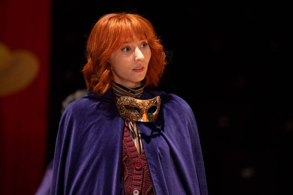 A woman with red hair wearing a blue cape and ornate mask stands on stage.