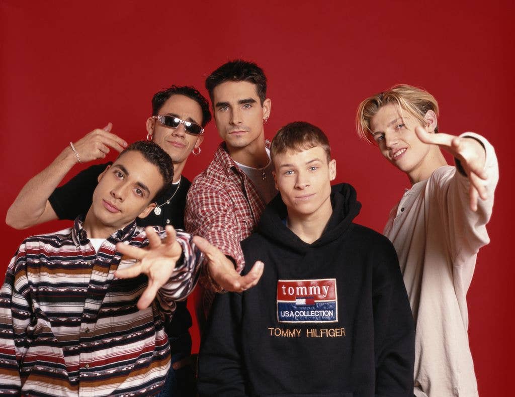 Five young men, including Brian Littrell, pose together against a red backdrop, several reaching towards the camera with their hands.