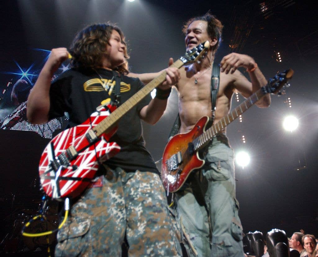 A young guitarist and an older shirtless guitarist play electric guitars together under bright stage lights, both smiling at each other.