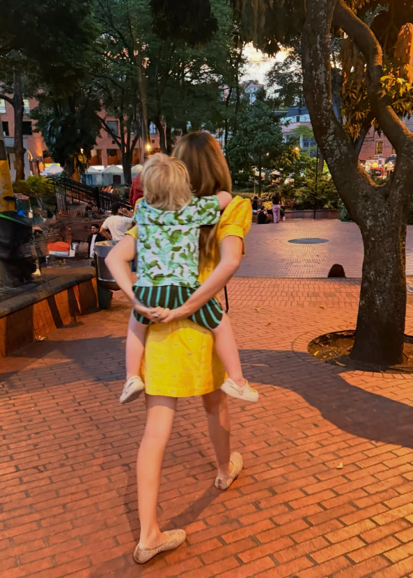 A woman in a yellow dress carries a child on her back in a park.