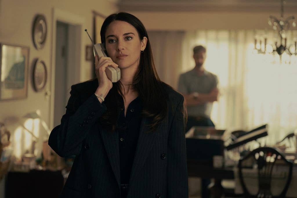 Shailene Woodley in a black suit, holding a phone, stands in a living room with a man in the background.