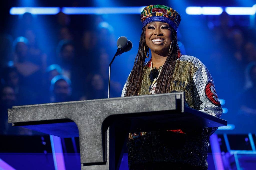 Missy Elliott smiles at a podium wearing a beaded crown and sequinned jacket, performing under blue stage lights
