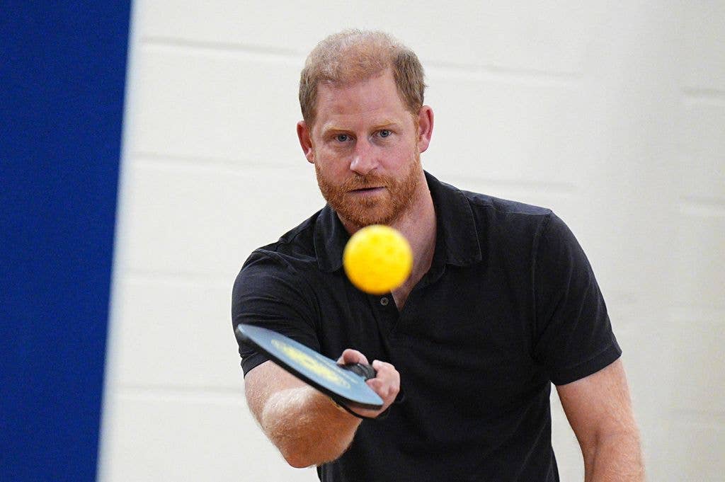Prince Harry, wearing a black polo shirt, focuses intently as he hits a yellow ball with a paddle during a pickleball game.
