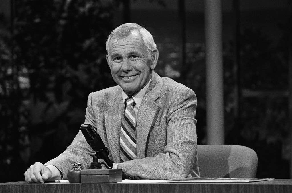 Black and white photo of Johnny Carson in a suit and striped tie, seated at his talk show desk and smiling, with a microphone in front of him.