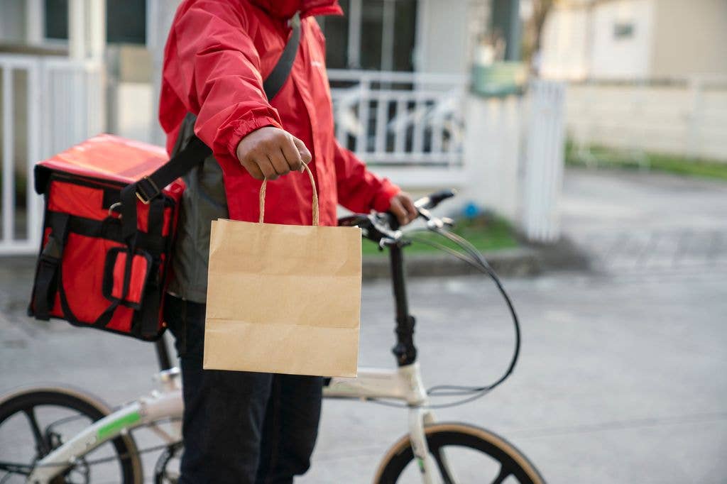A person in a red jacket stands beside a bicycle with a red insulated delivery box, holding out a brown paper bag by its handles.