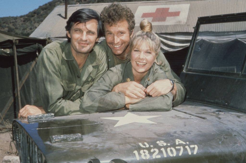 Three smiling actors in army fatigues lean over the bonnet of a military jeep in front of a tent marked with a red cross.