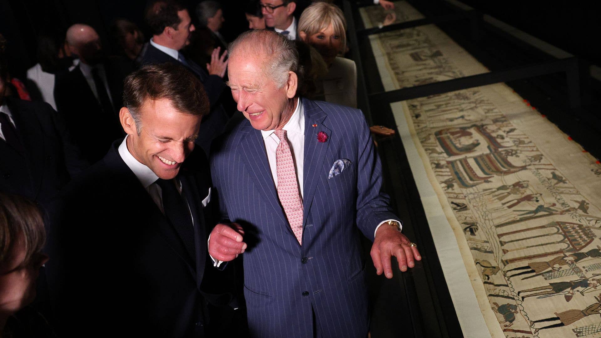 King Charles in a blue suit and pink tie, smiles with another man at an exhibit.