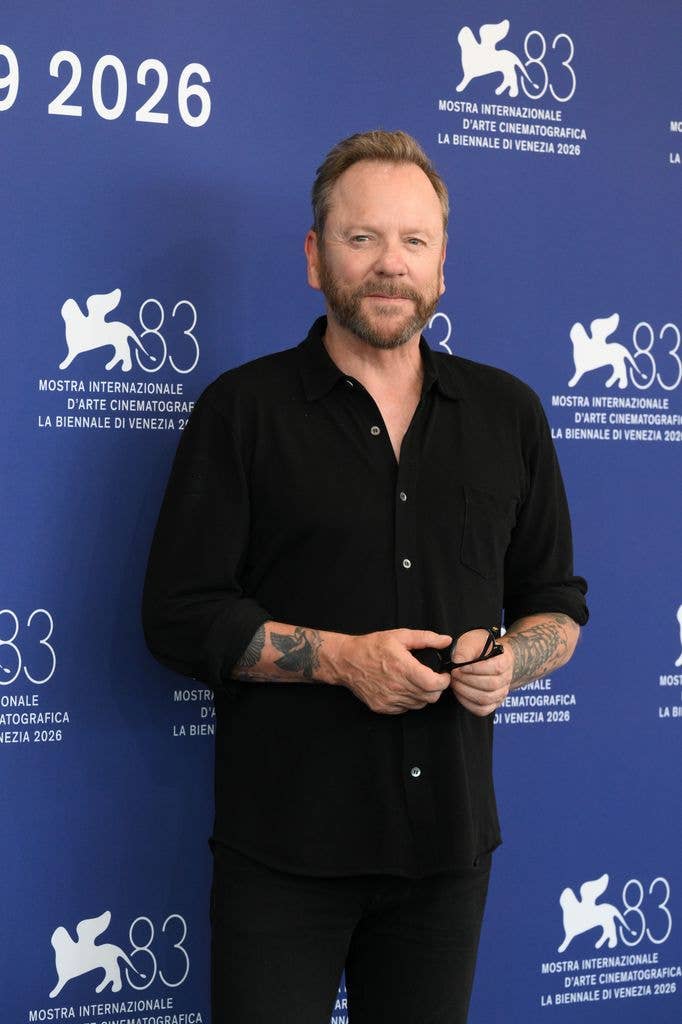 Kiefer Sutherland, wearing a black shirt and holding his glasses, poses in front of a blue Venice Film Festival backdrop.